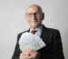 Senior Man In A Suit With Glasses Holding Us Dollars, Symbolizing Wealth And Savings.